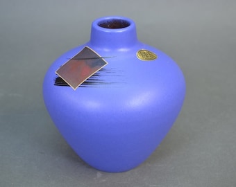 Può includere: Un piccolo vaso in ceramica blu con un design geometrico. Il vaso ha un design astratto nero e marrone su un lato e un adesivo dorato sull'altro.