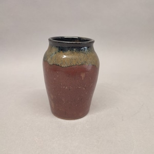 Puede incluir: Un pequeño jarrón de cerámica marrón con un esmalte negro y marrón. El jarrón tiene un cuerpo redondeado y un labio ligeramente acampanado.