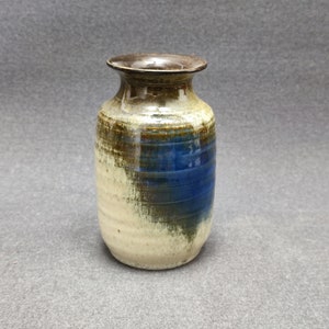 Puede incluir: Un pequeño jarrón de cerámica con un esmalte marrón, azul y blanco. El jarrón tiene un cuerpo redondeado y un cuello estrecho.
