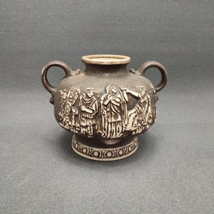 May include: A small brown ceramic vase with two handles and a white design of people and trees. The vase has a white base with a decorative pattern.