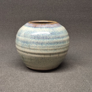 May include: A small, round ceramic vase with a light blue and white glaze. The vase has a brown rim and a subtle, striped pattern.