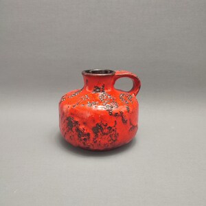 Könnte beinhalten: Eine kleine, rote Keramikvase mit einer strukturierten Oberfläche und einem schwarzen Henkel. Die Vase hat einen runden Körper und eine schmale Öffnung.
