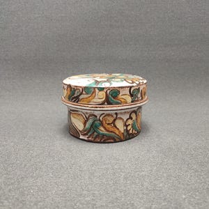 Puede incluir: Una pequeña caja redonda de cerámica con tapa. La caja está decorada con un patrón floral en tonos verdes, marrones y amarillos.