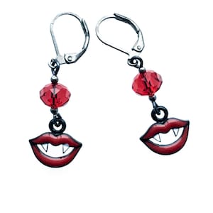 May include: A pair of silver leverback earrings. Each earring has a red crystal bead and a charm shaped like a red vampire mouth with white fangs. The earrings are set against a white background.
