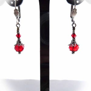 Puede incluir: Un par de pendientes de cristal rojo con detalles de filigrana plateada colgando de un soporte de pendientes negro.