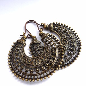 May include: A pair of antique brass-colored hoop earrings. The earrings feature intricate filigree patterns and small beaded accents along the edges. The ear wires are a darker bronze color. The earrings are displayed on a white background.