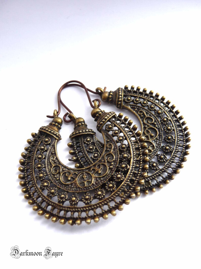 May include: A pair of antique brass-colored hoop earrings. The earrings feature intricate filigree patterns and small beaded accents along the edges. The ear wires are a darker bronze color. The earrings are displayed on a white background.