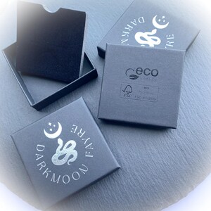 May include: Four black jewelry boxes with silver text and a snake and moon logo. One box is open, revealing a black interior. The boxes are on a dark gray surface. The text on the boxes reads "DARKMOON FAYRE".
