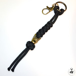 Peut inclure: Porte-clés en paracorde noir avec un charme tête de chat doré et un anneau de clé en laiton.