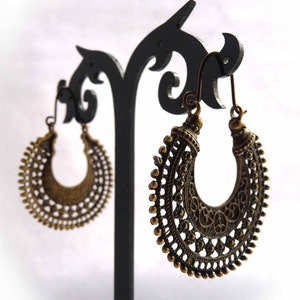 Puede incluir: Pendientes de aro de bronce ornamentados con intrincados detalles de filigrana y cuentas. Los pendientes se exhiben en un soporte negro sobre un fondo blanco. Los pendientes tienen una estética vintage o bohemia.