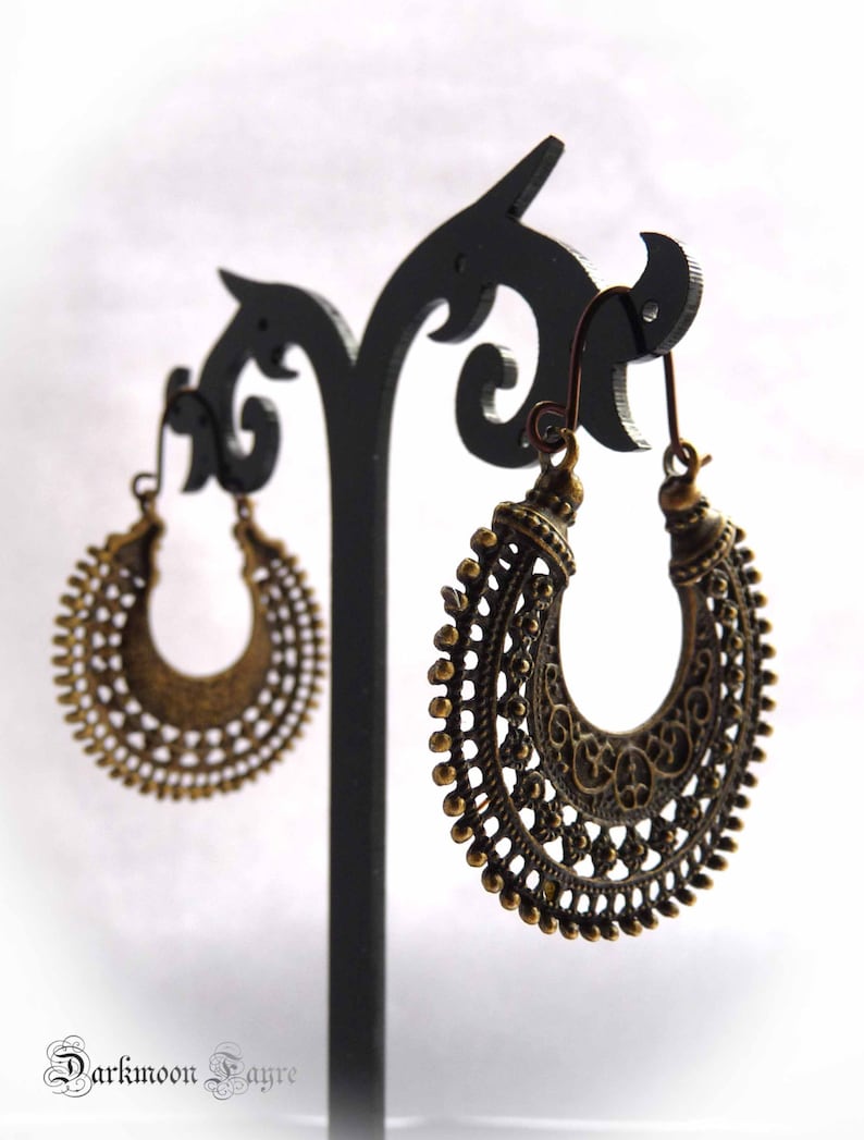 May include: Ornate bronze-colored hoop earrings with intricate filigree and beaded detailing. The earrings are displayed on a black earring stand against a white background. The earrings have a vintage or bohemian aesthetic.