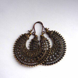 May include: A pair of antique brass-colored hoop earrings with intricate filigree and floral detailing. The earrings have a crescent shape with a decorative top and a hook closure. The earrings are displayed on a white background.