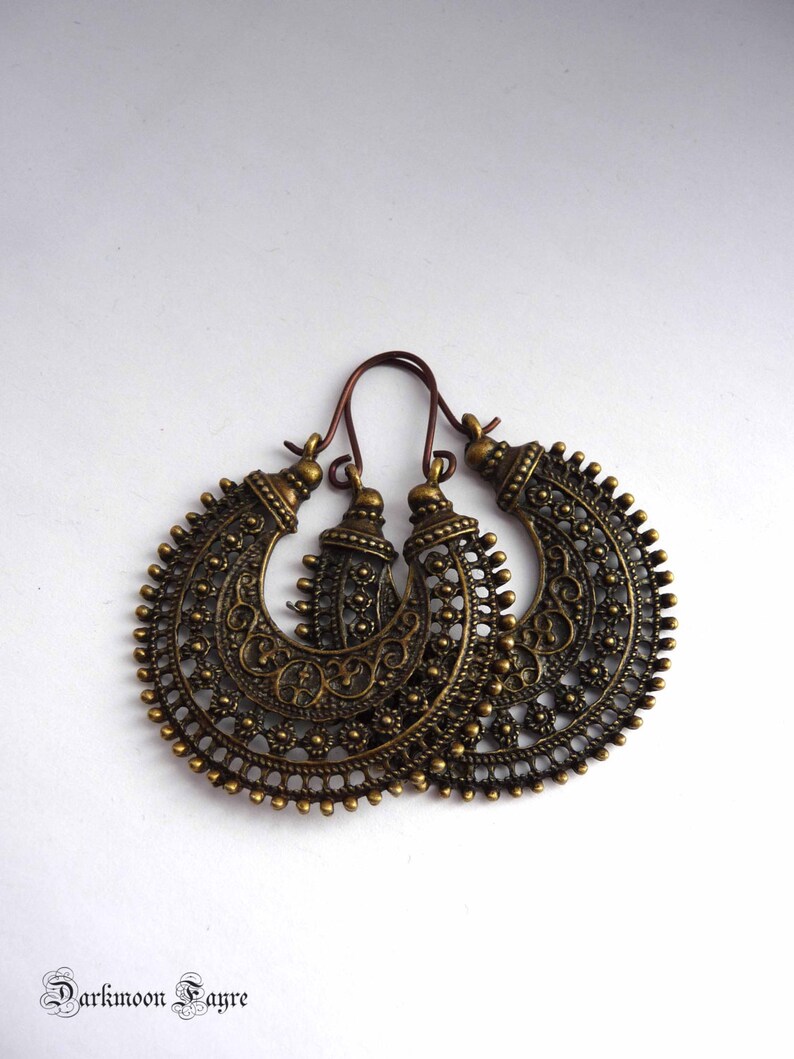 May include: A pair of antique brass-colored hoop earrings with intricate filigree and floral detailing. The earrings have a crescent shape with a decorative top and a hook closure. The earrings are displayed on a white background.