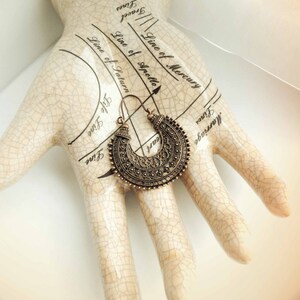 May include: A bronze-colored, ornate earring with a crescent shape is displayed on a ceramic hand with palmistry markings. The earring features intricate detailing and a hook closure. The image includes the text "Darkmoon Fayre".