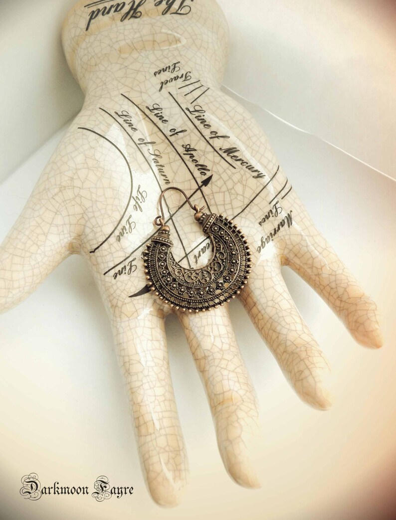 May include: A bronze-colored, ornate earring with a crescent shape is displayed on a ceramic hand with palmistry markings. The earring features intricate detailing and a hook closure. The image includes the text "Darkmoon Fayre".