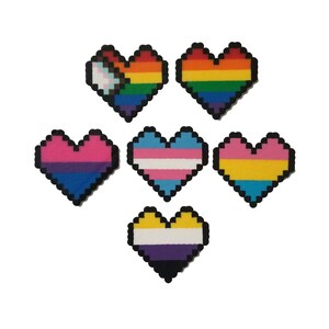 May include: Pixelated heart-shaped designs in various colors representing LGBTQ+ pride flags. The hearts include the rainbow flag, transgender flag, bisexual flag, pansexual flag, non-binary flag, and a pride flag with a white, pink, and blue design.