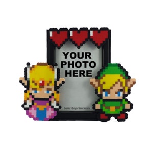 May include: A black photo frame with pixelated characters of a princess and a hero, with the text "YOUR PHOTO HERE". The frame has three red heart pixel art at the top. The characters are in yellow, pink, green, and blue.