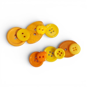 May include: Two sets of decorative buttons in shades of yellow and orange. Each set includes various sizes and shapes, including round and apple-shaped buttons. The buttons are arranged on a white background.
