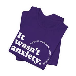 May include: A folded purple t-shirt with white text that reads "It wasn't anxiety." A semi-circle of text around the center reads "Superior Mesenteric Artery Syndrome."