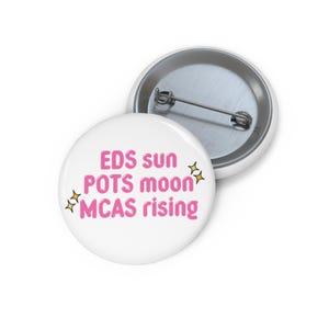 May include: White button pin with the text "EDS sun POTS moon MCAS rising" in pink, with yellow star accents. The pin is round and has a metal backing with a pin clasp.