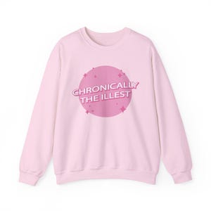 Op de afbeelding: Een lichtroze sweatshirt met de tekst "Chronically The Illest" in een roze cirkel. De tekst is in een retro lettertype, met sterren rond de cirkel. De sweatshirt heeft een ronde hals.