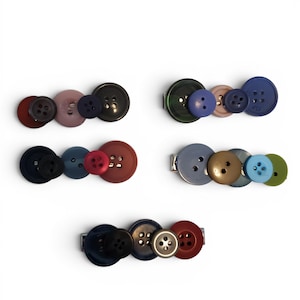 May include: A collection of button hair clips in various colors, including red, pink, blue, green, and black. Each clip features an assortment of buttons attached to a silver-tone metal barrette.