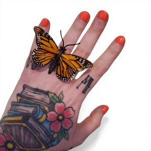 May include: A monarch butterfly figurine with orange and black wings, detailed with white spots, rests on a hand. The butterfly's body is black. The hand has orange nail polish and tattoos of books, flowers, and other designs.