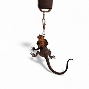 Könnte beinhalten: Ein Metall-Schlüsselanhänger mit einem Kragenechsen-Design. Die Echse ist braun mit einem beigefarbenen Kragen und ist an einem Metallverschluss und Schlüsselring befestigt. Der Schlüsselanhänger hängt an einem dunkelbraunen Riemen vor weißem Hintergrund.