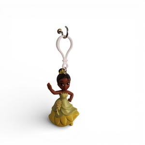 May include: A small, plastic figurine of a princess in a green and yellow dress. The princess has dark skin and a bun hairstyle. The figurine is attached to a white clip with a silver-colored metal ring.