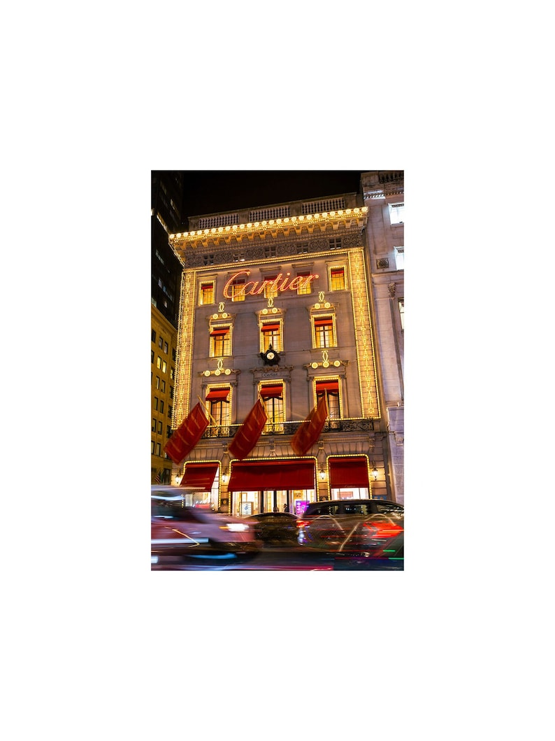 May include: A large, ornate building with the word 'Cartier' in red letters above the entrance. The building is lit up with yellow string lights and has red awnings over the windows. There are cars driving by in the street in front of the building.