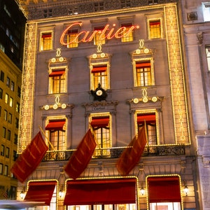 May include: A storefront with the word 'Cartier' in red neon letters above the entrance. The building is decorated with white string lights and red flags. There are two cars in the foreground, one is blurred and the other is partially visible.