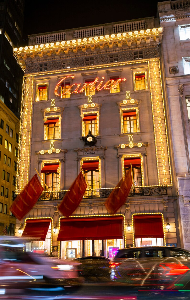 May include: A storefront with the word 'Cartier' in red neon letters above the entrance. The building is decorated with white string lights and red flags. There are two cars in the foreground, one is blurred and the other is partially visible.