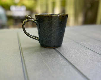 Pode incluir: Uma caneca de cerâmica azul escura com um esmalte salpicado. A caneca tem uma alça arredondada e uma base larga.