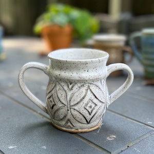 May include: White ceramic mug with two handles and a geometric pattern. The mug has a speckled glaze.