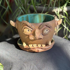 May include: A handcrafted ceramic plant pot with a face design. The brown pot has a turquoise interior and a matching saucer. The face has exaggerated features, including large eyes, a nose, and teeth. Small horns are on the sides.