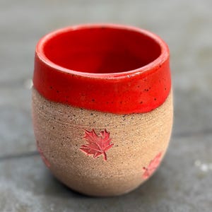 Puede incluir: Una taza de cerámica hecha a mano con interior y borde rojos. El exterior es de color canela con un acabado texturizado y diseños de hojas de arce rojas en relieve. La taza está diseñada para bebidas y es adecuada para uso doméstico.