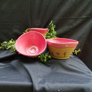May include: Three pink ceramic bowls with a brown rim. The largest bowl has a brown rim and a design of birds on the bottom. The bowls are on a black fabric surface.