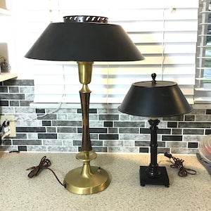 May include: Two table lamps with black shades. The larger lamp has a gold base with a wood-like design and a black shade with a decorative metal trim. The smaller lamp has a black base and a black shade.