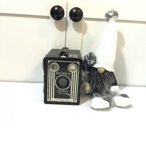 May include: A black and silver vintage Brownie Target Six-20 camera with a white and gray gnome figurine. The camera has a black leather strap and the gnome has a white hat and a gray beard.