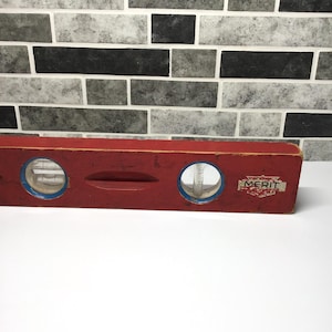 May include: A vintage red carpenter's level with two circular bubble viewers and a central oval indentation. The level has blue accents around the viewers and the word "MERIT" is printed on a small label. The tool is made of wood and shows signs of wear.