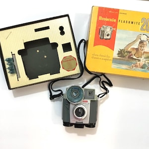 May include: A vintage Kodak Brownie Flashmite 20 camera with a flash attachment and a black strap. The camera is silver and black with a red Kodak logo. The camera is in its original box, which is yellow with red and black text. The box has a picture of a woman in a swimming pool.