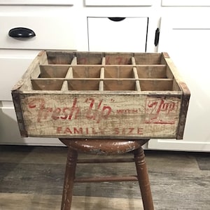 May include: Vintage wooden crate with compartments, featuring the "Fresh Up" logo in red lettering. The crate is weathered with metal reinforcements on the corners. It is labeled "Family Size" and has the 7 Up logo. The crate is sitting on a wooden stool.