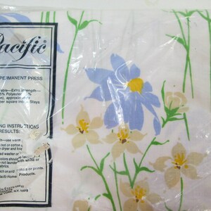 May include: A white sheet with a floral pattern featuring blue, yellow, and green flowers. The sheet is packaged in plastic and has a label that reads "Pacific" and "No Iron/Permanent Press".