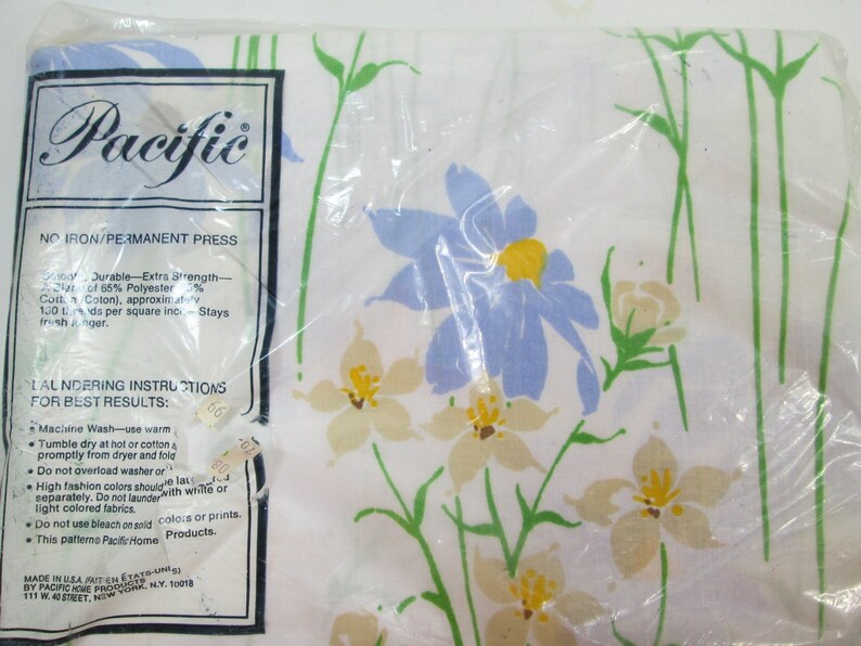 May include: A white sheet with a floral pattern featuring blue, yellow, and green flowers. The sheet is packaged in plastic and has a label that reads "Pacific" and "No Iron/Permanent Press".