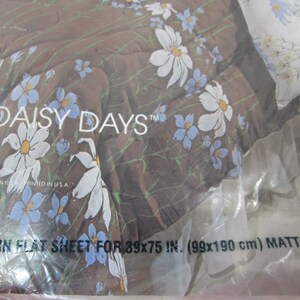May include: A close-up of a brown and white twin flat sheet with a daisy print. The sheet is packaged in plastic and has the text "Daisy Days" and "Twin Flat Sheet for 39x75 in. (99x190 cm) Mattress-F" printed on it.