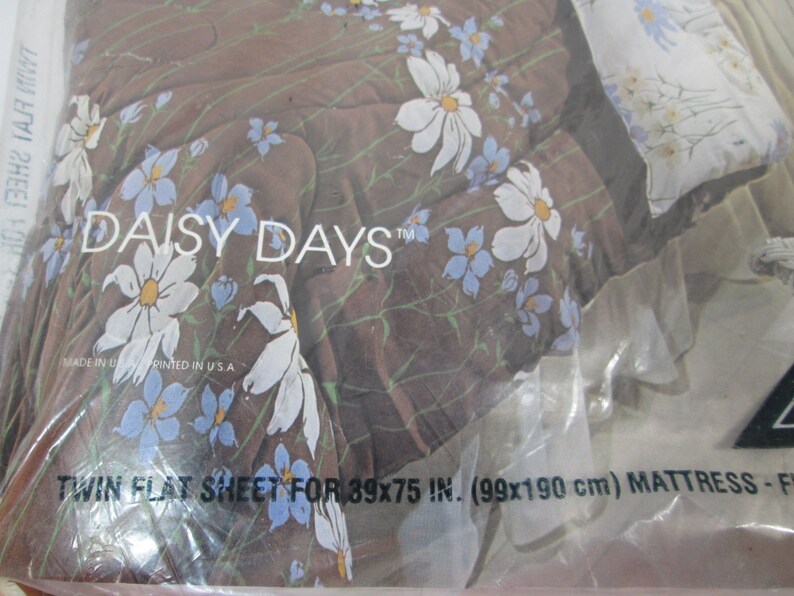 May include: A close-up of a brown and white twin flat sheet with a daisy print. The sheet is packaged in plastic and has the text "Daisy Days" and "Twin Flat Sheet for 39x75 in. (99x190 cm) Mattress-F" printed on it.