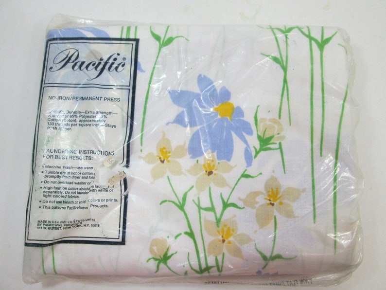 May include: A white sheet with a floral pattern featuring blue and yellow flowers. The sheet is packaged in plastic and has a label that reads "Pacific" and "No Iron/Permanent Press".
