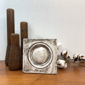 May include: A white square plaque with a raised, circular design in the center. The plaque has a distressed, aged look with a gray wash over the white paint. The plaque is sitting on a wooden surface.