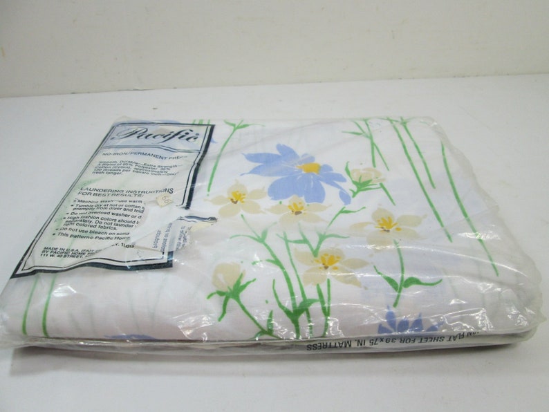 May include: A white flat sheet with a floral pattern in shades of blue, yellow, and green. The sheet is packaged in plastic and labeled "Pacific No Iron Permanent Press" with laundering instructions. The label also states "Flat Sheet for 39x75 in. Mattress".