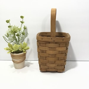 May include: A small, woven basket with a wooden handle. The basket is light brown and has a rectangular shape. Next to the basket is a small potted plant with green and yellow flowers.
